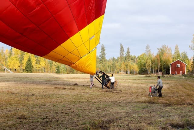 Nederdel av ballong som reser sig under fyllning på slagen gulnad åker med höstfärgad skog i bakgrunden